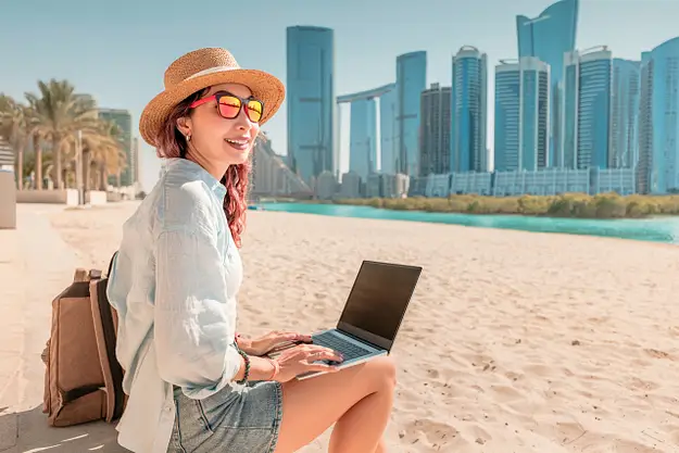 Woman using laptop on Abu Dhabi beach with skyline representing residency options and lifestyle benefits of property investment