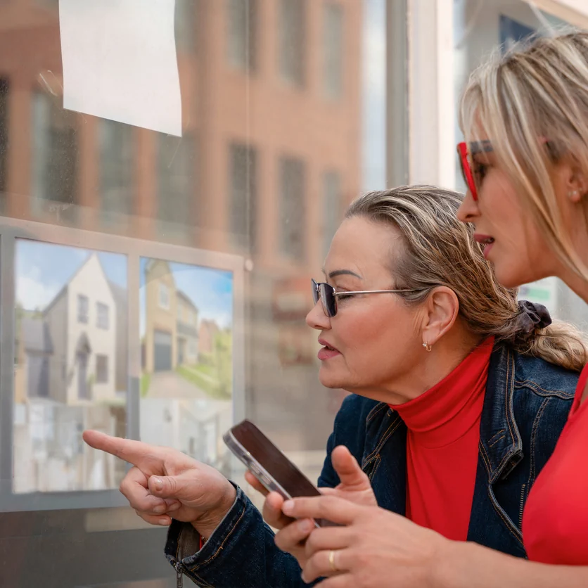 Two women reviewing property listings in estate agent window discussing UK property investment opportunities and housing options